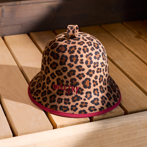 A leopard print sauna hat with embroidered 'Sweltry' logo, displayed on a light wood sauna bench.