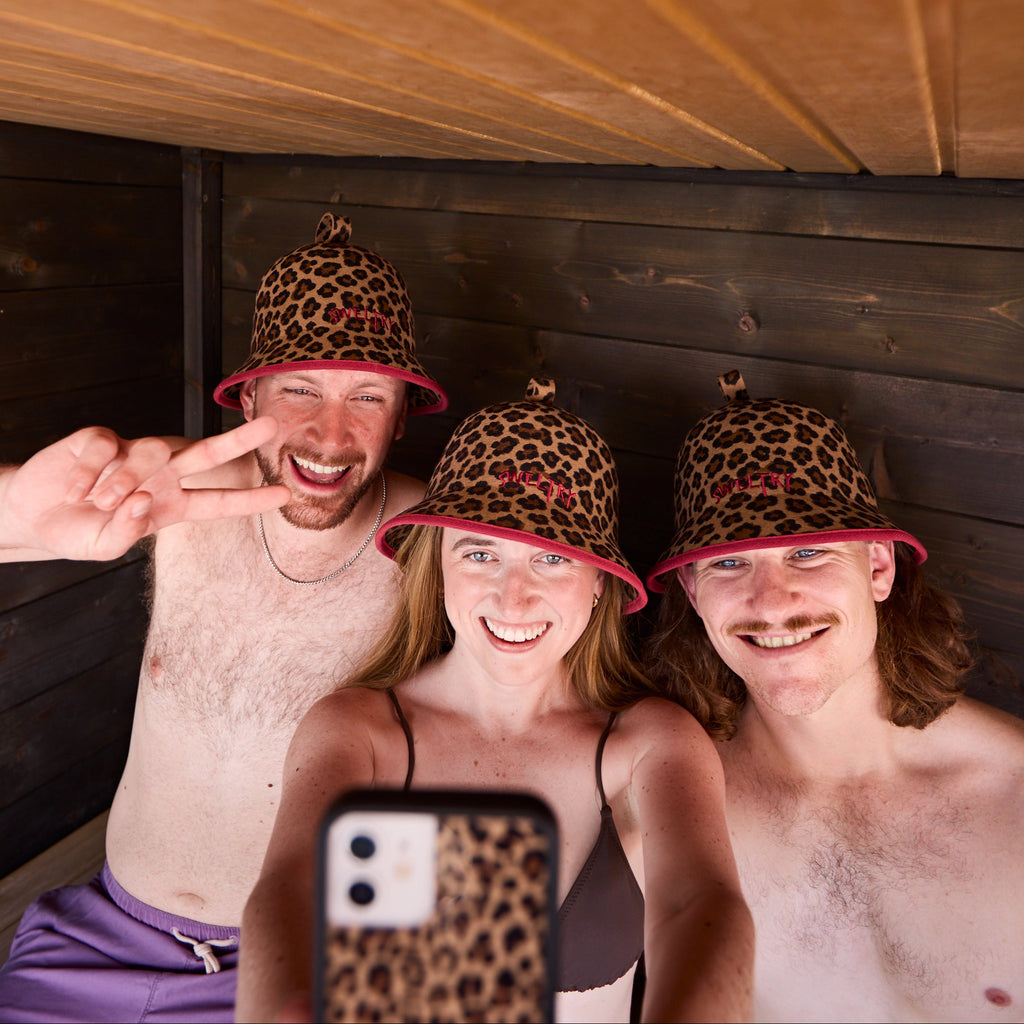 Three people taking a selfie inside a wooden sauna wearing matching leopard-print sauna caps.