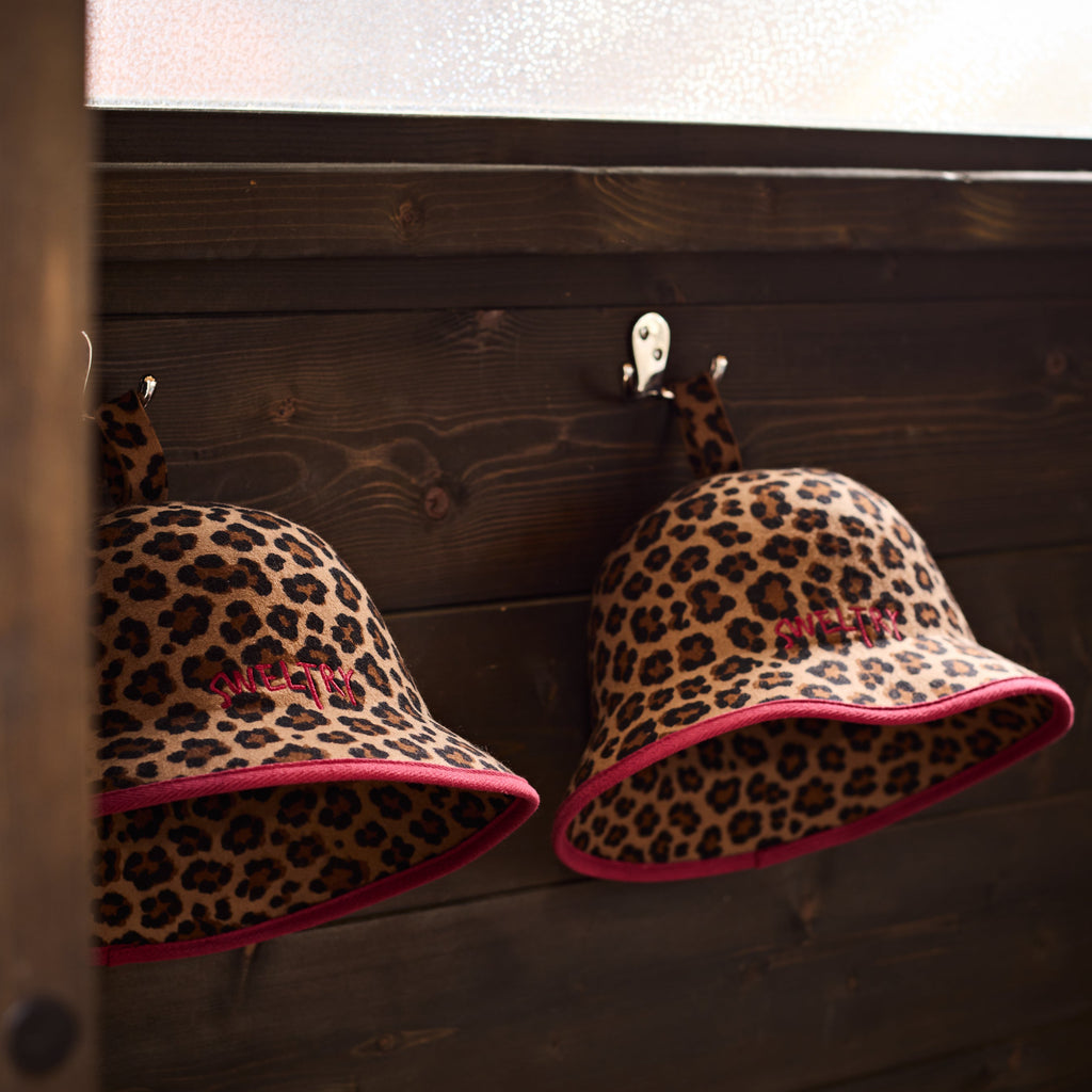 Two leopard-print sauna bucket hats hanging on hooks against a dark wood sauna wall.