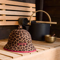 A leopard-print bucket hat with burgundy binding and embroidered Sweltry logo, displayed on a light wood sauna bench next to traditional sauna accessories.