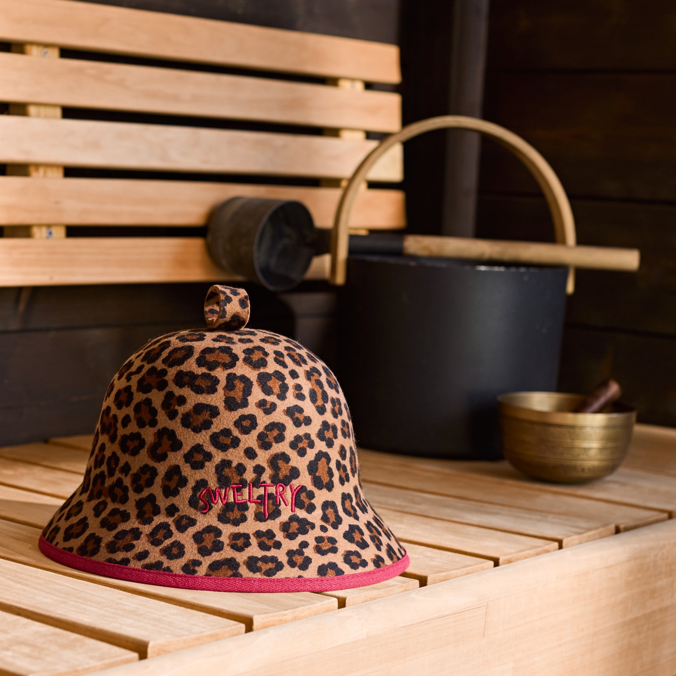 A leopard-print bucket hat with burgundy binding and embroidered Sweltry logo, displayed on a light wood sauna bench next to traditional sauna accessories.