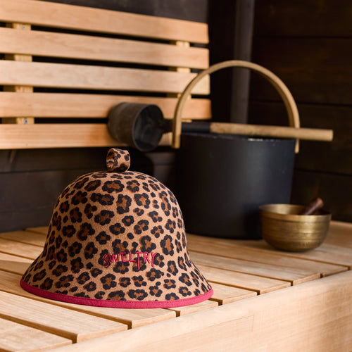 A leopard-print bucket hat with burgundy binding and embroidered Sweltry logo, displayed on a light wood sauna bench next to traditional sauna accessories.