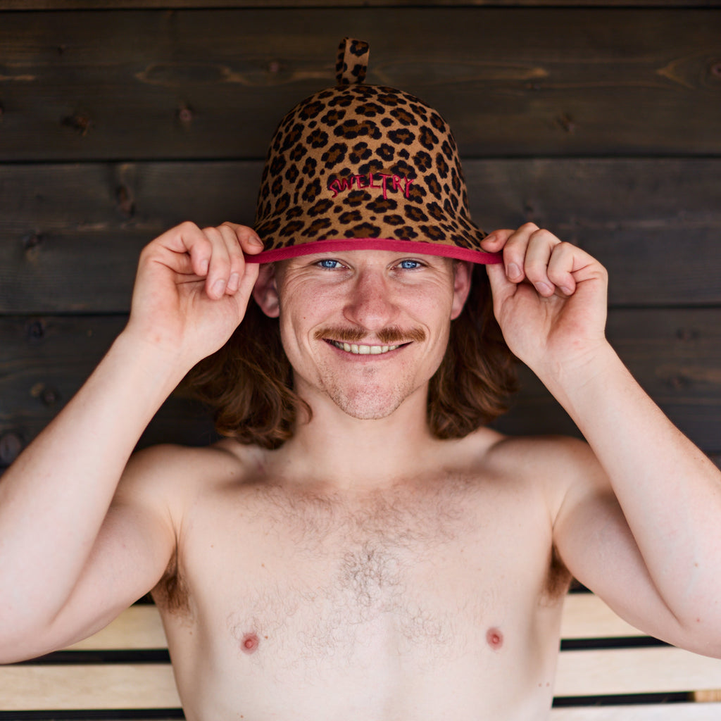 A shirtless man with a mustache pulls on a leopard-print sauna hat.