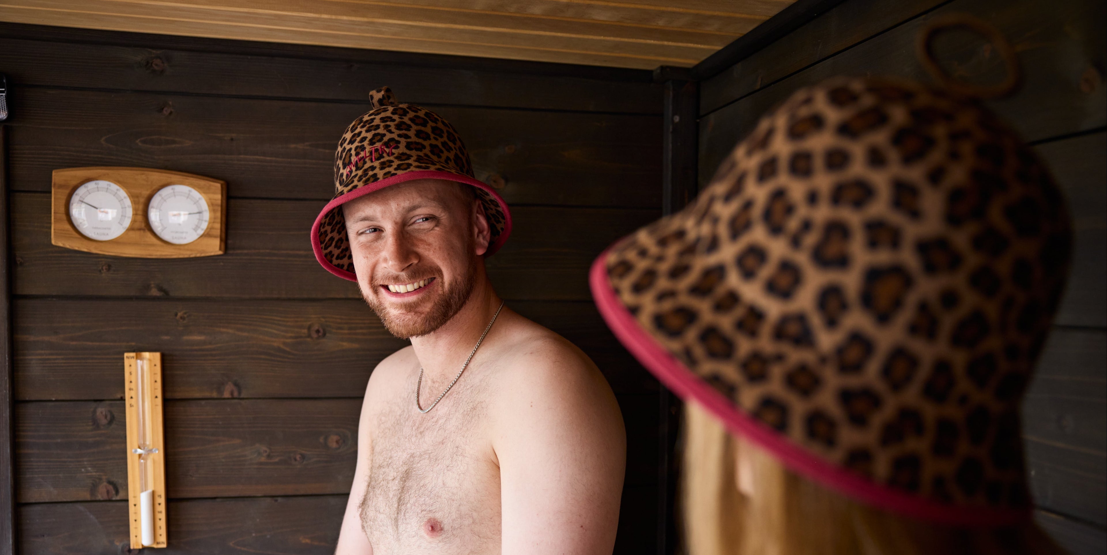 A man in a leopard-print sauna hat with a burgundy trim smiles inside a wooden sauna, while another person wearing an identical hat is shown from behind in the foreground.